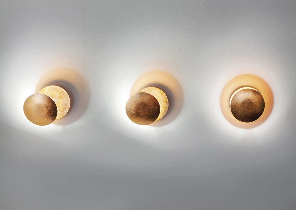 Three Clérachic wall lights in a row on a pale wall, each in a different gold or bronze tone, all lit with a soft glow.