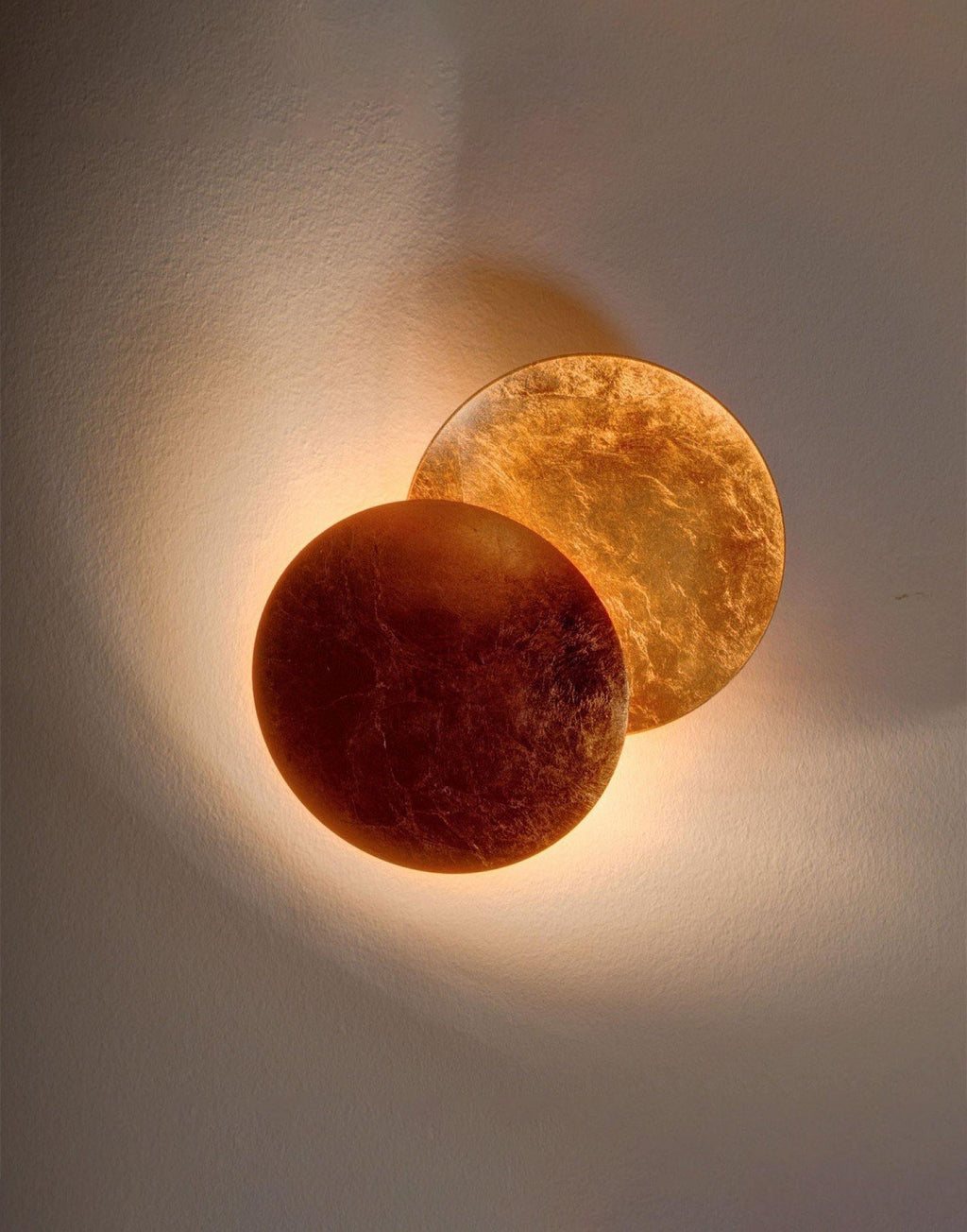 Upright view of this Clérachic wall light lit on a dark taupe wall, a deep bronze disc in front of a gold-leaf disc.