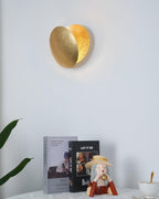 Gold Clérachic wall light on a white wall above a side table with books, a plant and a small figurine, its front disc tilted forward.