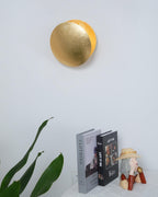 This Clérachic wall light in gold, seen from the front on a white wall above two stacked books and a leafy plant.