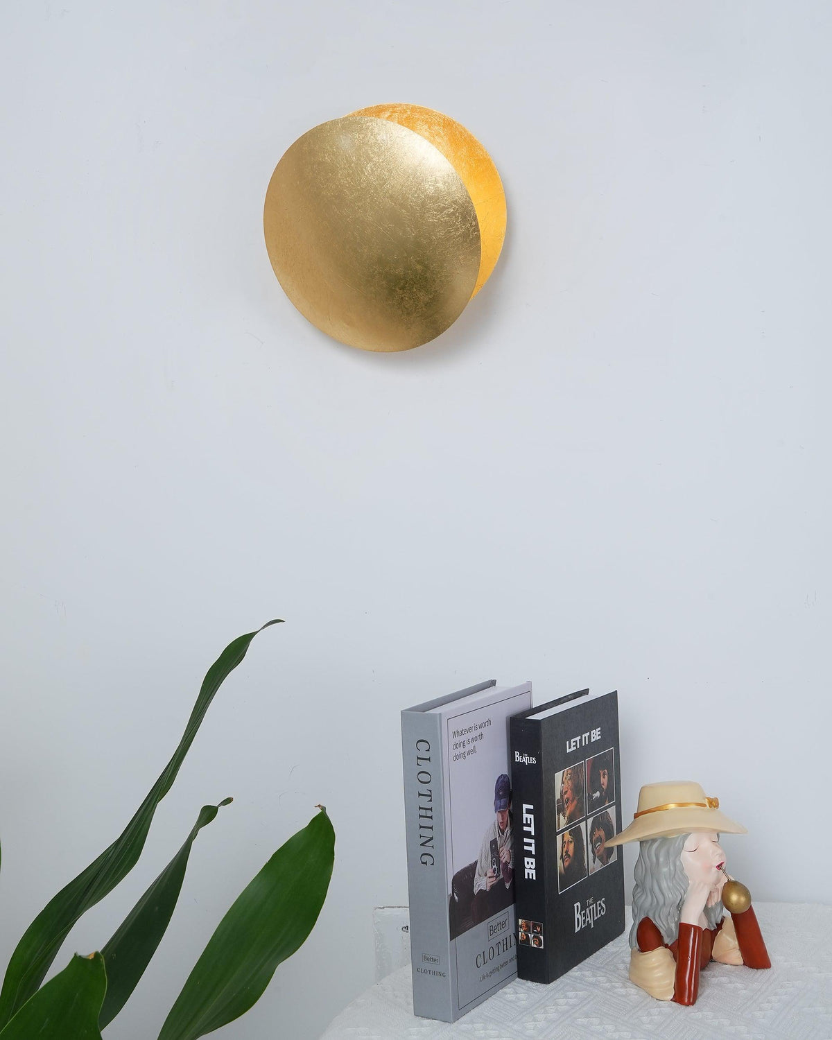 This Clérachic wall light in gold, seen from the front on a white wall above two stacked books and a leafy plant.