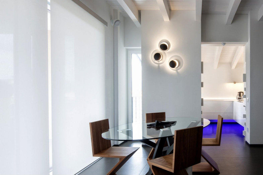 Three dark-finished Clérachic wall lights grouped on a white column in an open-plan dining room with a glass table.