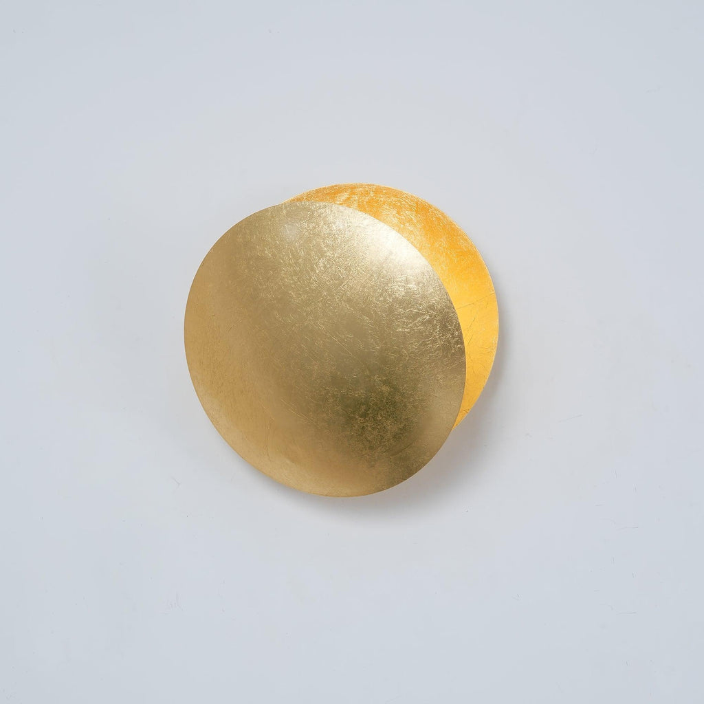 Gold Clérachic wall light on a pale grey-blue wall, its front disc angled so a brighter gold disc glows behind it.