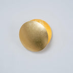 Gold Clérachic wall light on a pale grey-blue wall, its front disc angled so a brighter gold disc glows behind it.