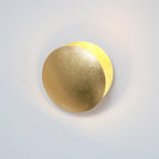 Gold-leaf Clérachic wall light with a soft light spilling from behind the front disc onto a pale wall.
