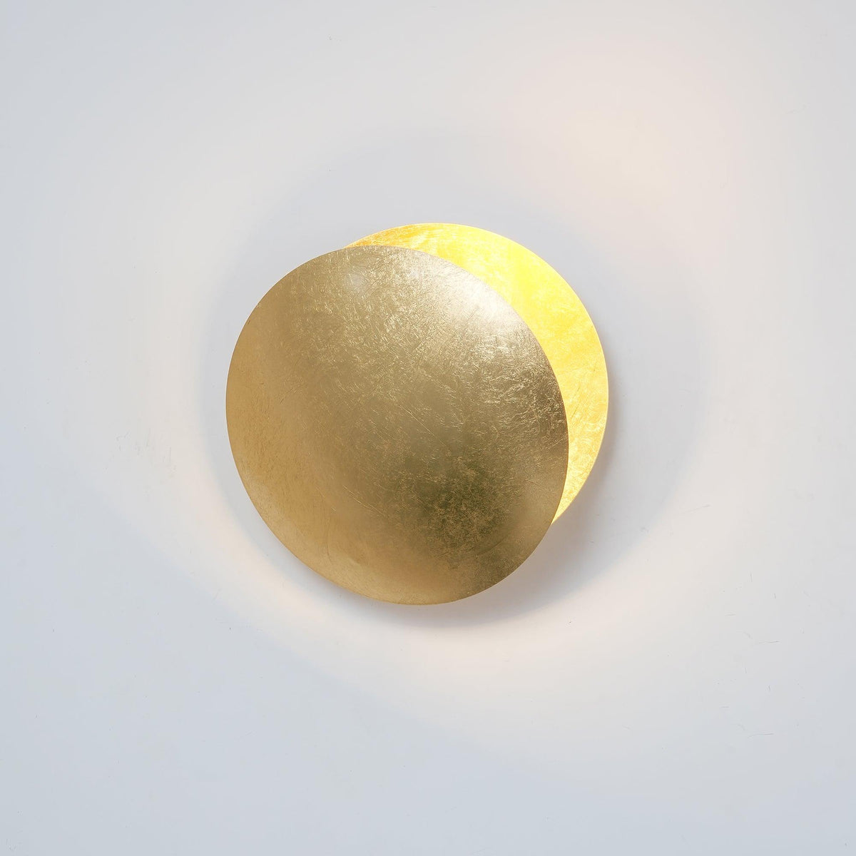 Gold-leaf Clérachic wall light with a soft light spilling from behind the front disc onto a pale wall.