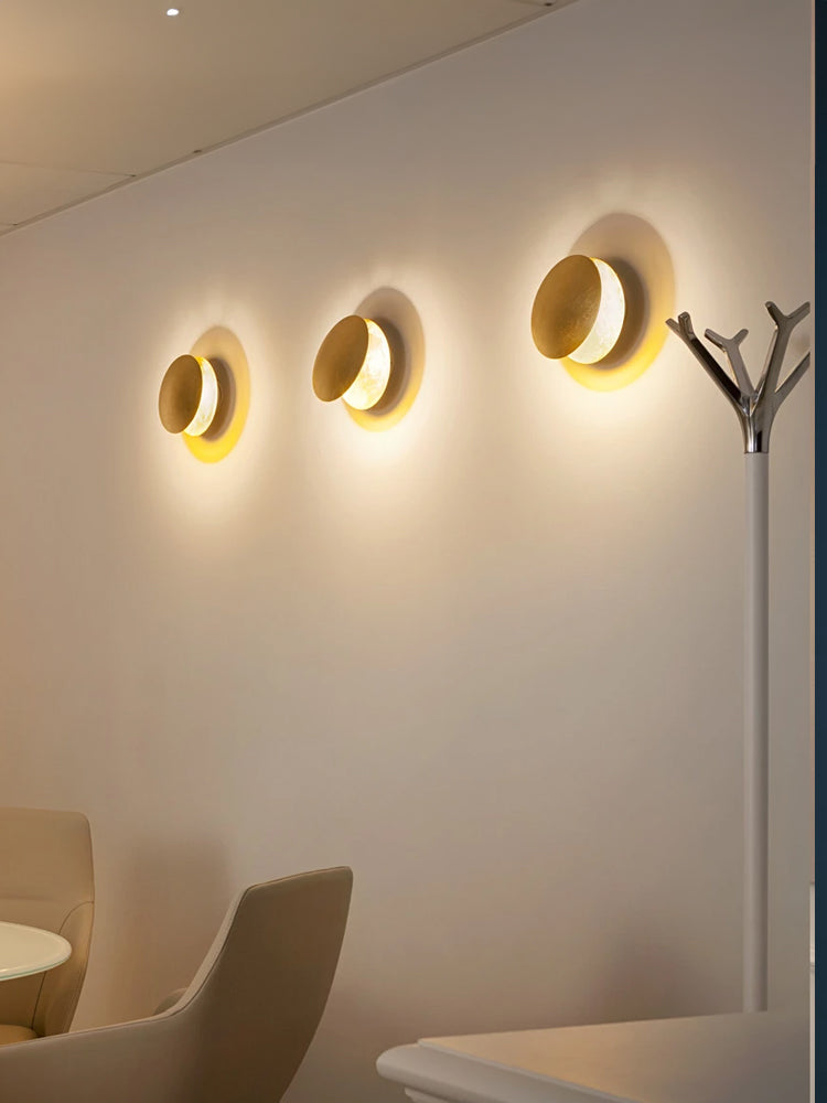 Three lit Clérachic wall lights in a row on a warm cream wall in a waiting area, beside a slim coat stand.