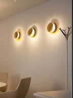 Three lit Clérachic wall lights in a row on a warm cream wall in a waiting area, beside a slim coat stand.