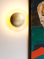 Tight view of a Clérachic wall light with a pale silvery disc and a ring of gold light, beside the edge of a framed painting.