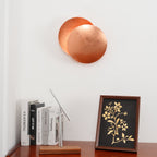 Copper Clérachic wall light on a white wall above a wooden sideboard with books, a slim silver lamp and a framed gold-leaf print.