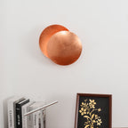 Closer view of this copper Clérachic wall light on a white wall, above a row of books and a framed floral print.