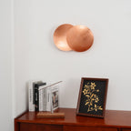 Wide view of a copper Clérachic wall light high on a white wall over a polished wood sideboard, books and a framed print.