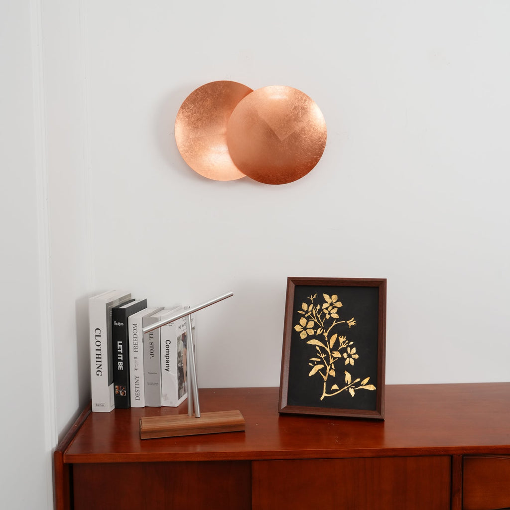 Copper Clérachic wall light hung over a dark-wood sideboard with books, a small desk lamp and a framed floral print.