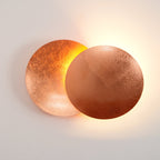 Lit copper Clérachic wall light on a white wall, the rear disc glowing orange and a soft light washing the surface around it.