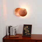 Copper Clérachic wall light switched on above a wooden sideboard, with a soft warm halo on the white wall behind.