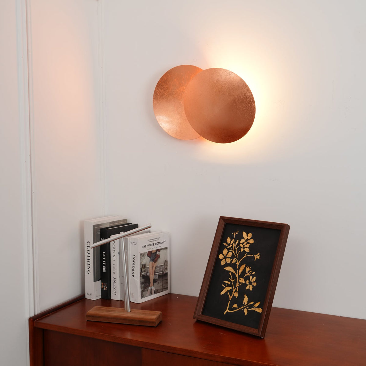 Lit copper Clérachic wall light on a white wall over a sideboard, books and a framed print, with a gentle glow around the discs.