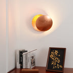 Clérachic wall light in copper with its rear disc glowing yellow-gold, on a white wall above a sideboard, books and a framed print.