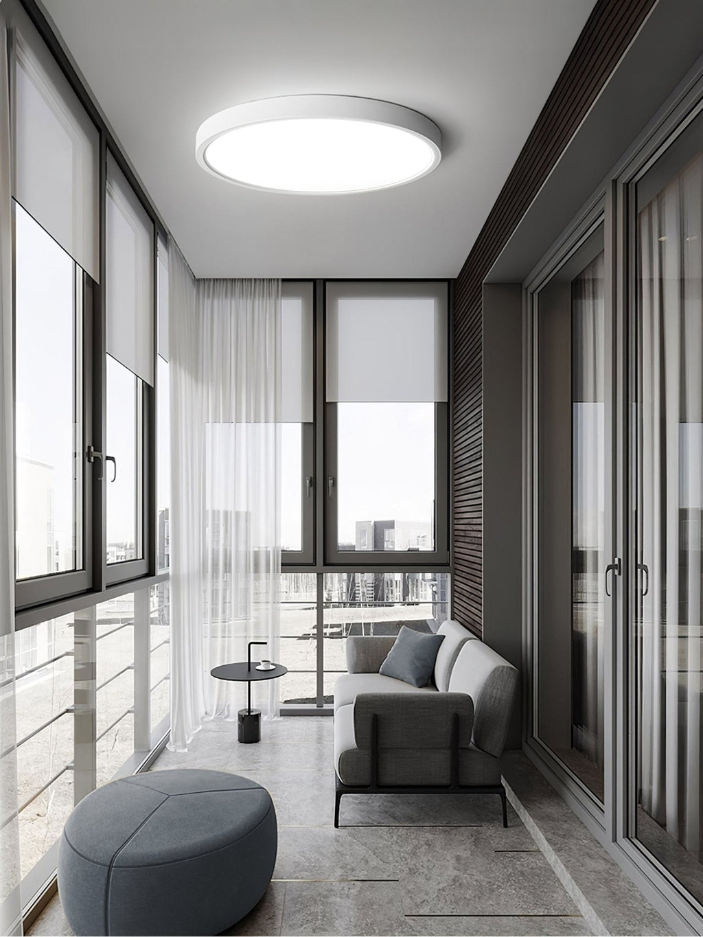 In a glazed sunroom with sheer curtains, a white Clérachic round ceiling light hangs over a grey sofa and a round blue-grey pouffe.