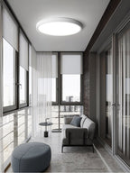 In a glazed sunroom with sheer curtains, a white Clérachic round ceiling light hangs over a grey sofa and a round blue-grey pouffe.