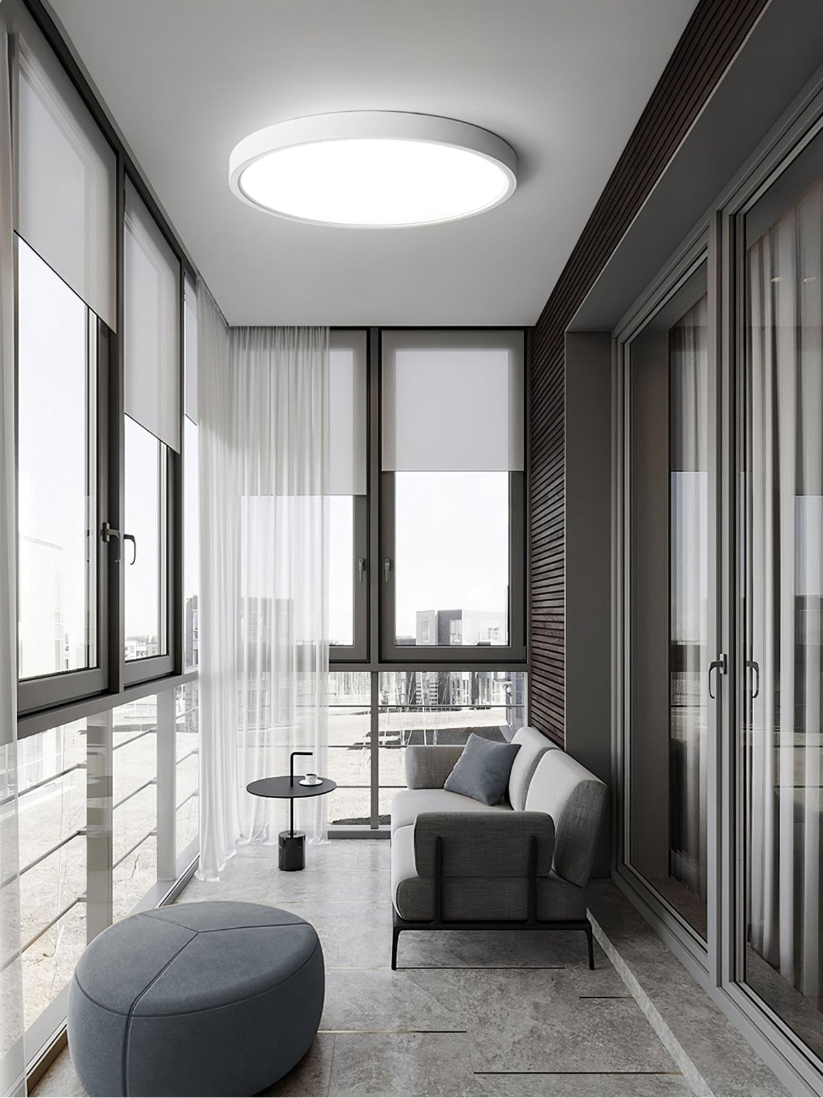 In a glazed sunroom with sheer curtains, a white Clérachic round ceiling light hangs over a grey sofa and a round blue-grey pouffe.