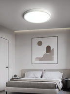 A white Clérachic round ceiling light glows above a grey bed with white pillows, beneath a framed abstract print.