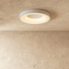 Clérachic white round ceiling light in a slightly larger size, lit warm white over a plaster wall.