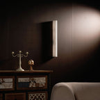 Unlit alabaster column sconce on a dark brown panelled wall, with a candelabra on a patterned cabinet below; this Clérachic wall light.
