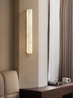 A Clérachic alabaster sconce glowing on a pale pillar beside heavy curtains, with a wooden shelf and a mug below.