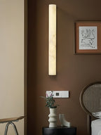 Tall alabaster column wall light lit above a small framed print and a rattan chair, on a warm brown wall; from Clérachic.