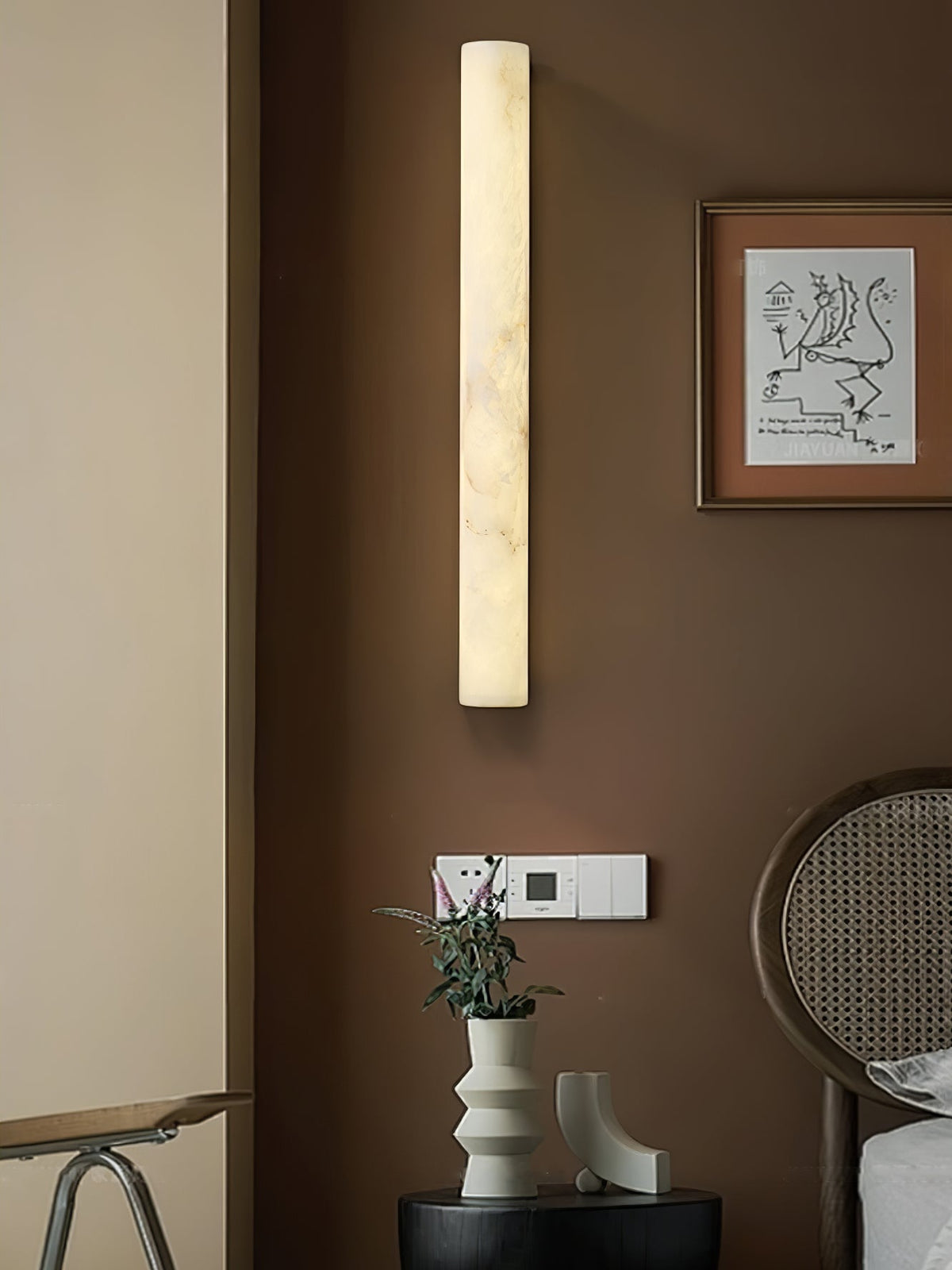 Tall alabaster column wall light lit above a small framed print and a rattan chair, on a warm brown wall; from Clérachic.