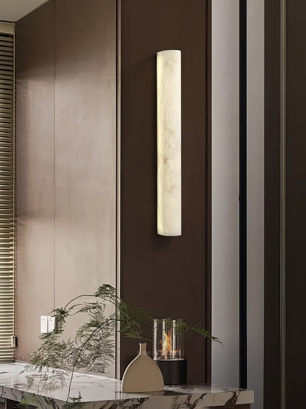 This Clérachic sconce glows on a brown panelled wall above a marble console, with a trailing plant and a glass candle holder.