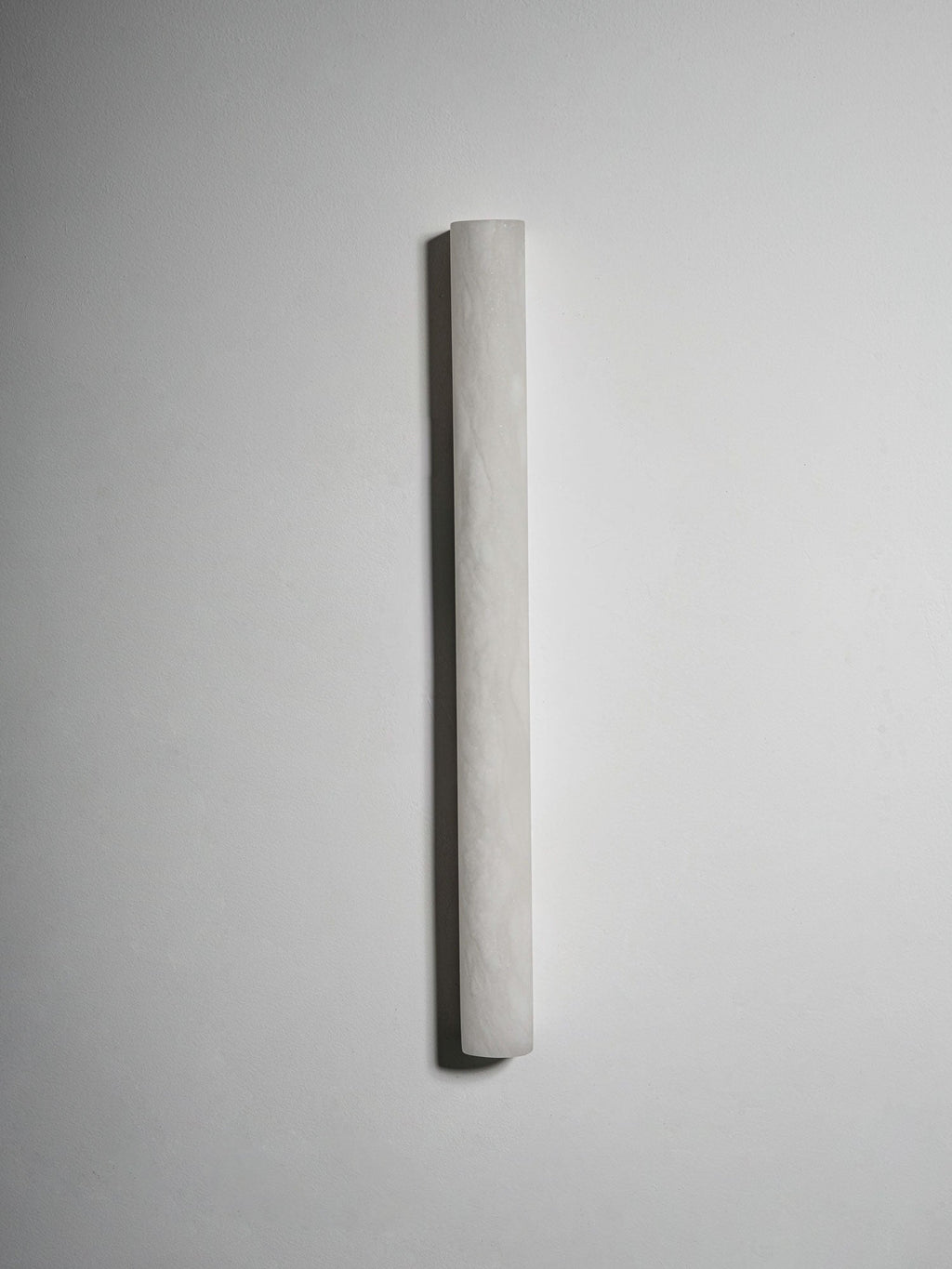 Unlit slim sconce on a grey wall, its pale alabaster surface showing a faint hand-finished texture; a Clérachic wall light.