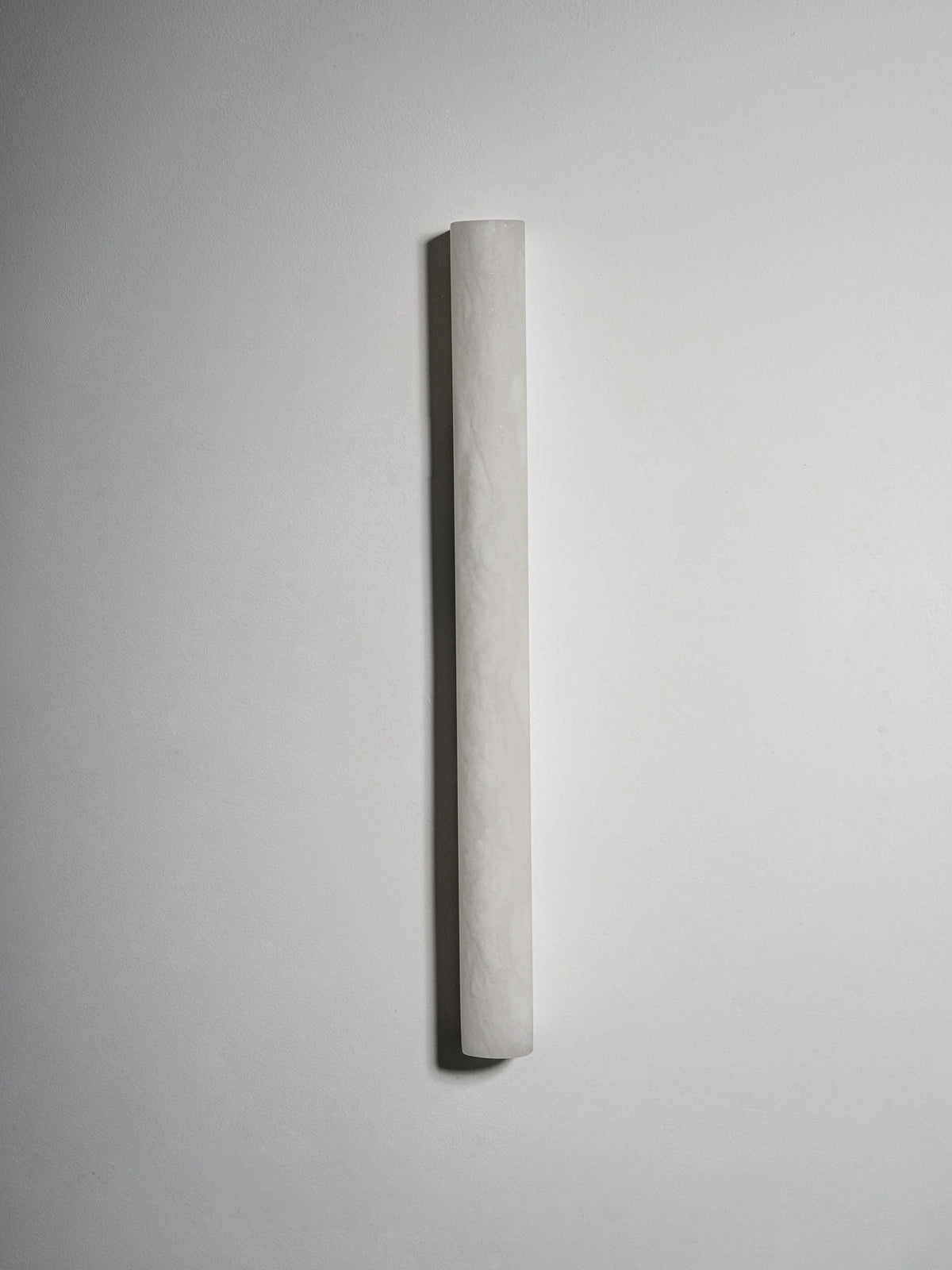 Unlit Clérachic sconce on a pale grey wall, its slim half-cylinder form in white alabaster with a faint rippled texture.