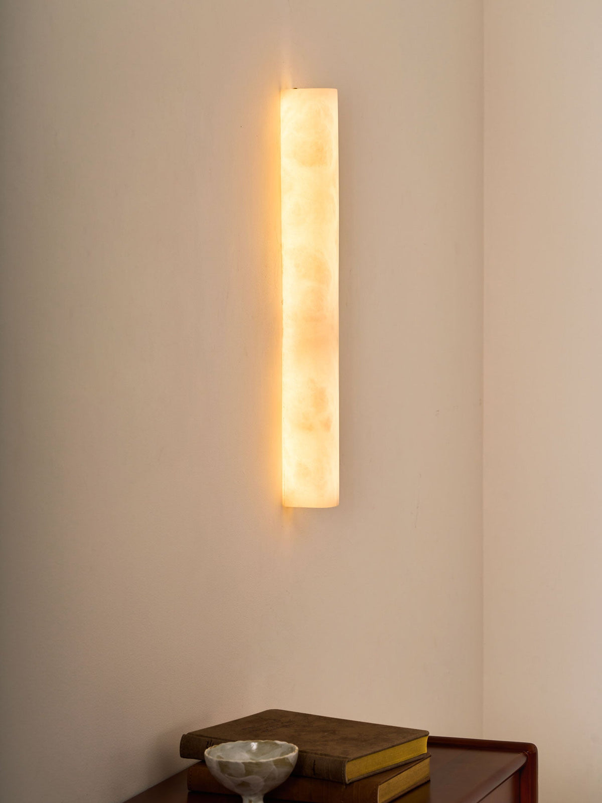 Warm-glowing alabaster sconce in a room corner above a wooden table with books and a small bowl; a Clérachic wall light.