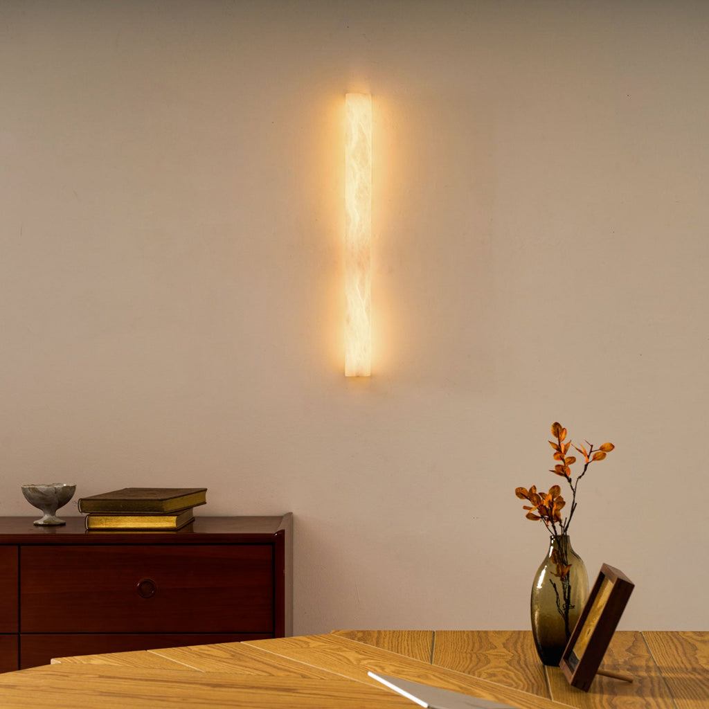 This Clérachic sconce glows warm on a cream wall above a wooden desk with a vase of dried stems and a framed picture.