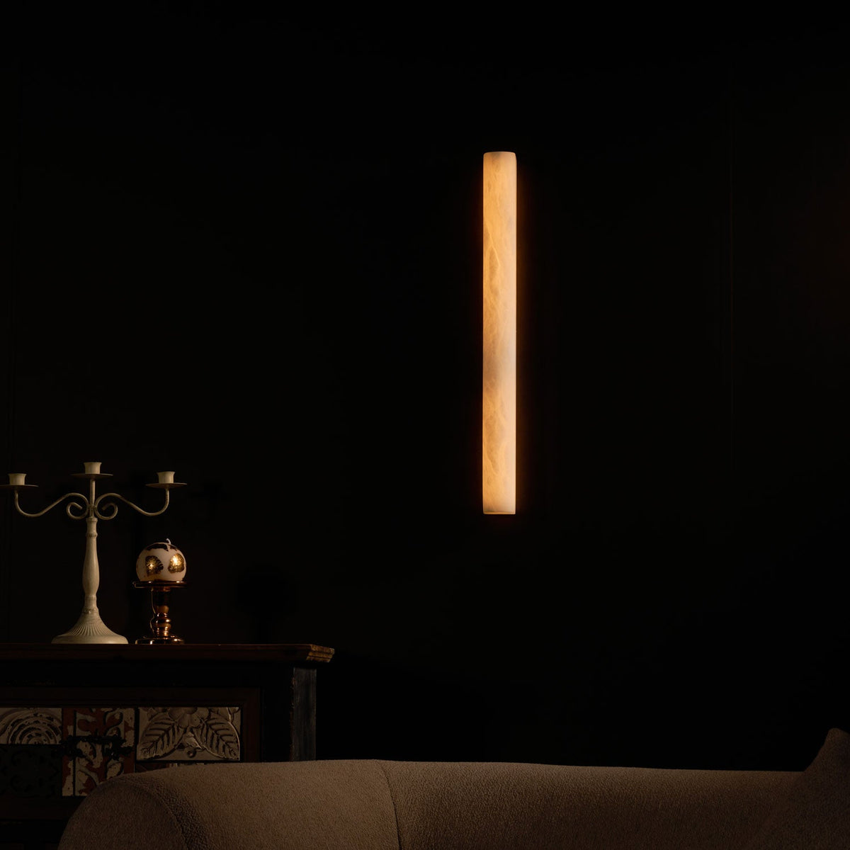 Narrow amber strip of light on a very dark wall above a candelabra and a patterned cabinet; the sconce in a night scene from Clérachic.