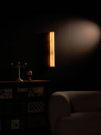 Distant view of the lit sconce glowing amber high on a dark wall, above a cabinet, a candelabra and a rounded armchair; Clérachic.