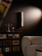 Unlit alabaster sconce on a dark wall lit by a soft spotlight, above a cabinet and a rounded cream armchair; from Clérachic.