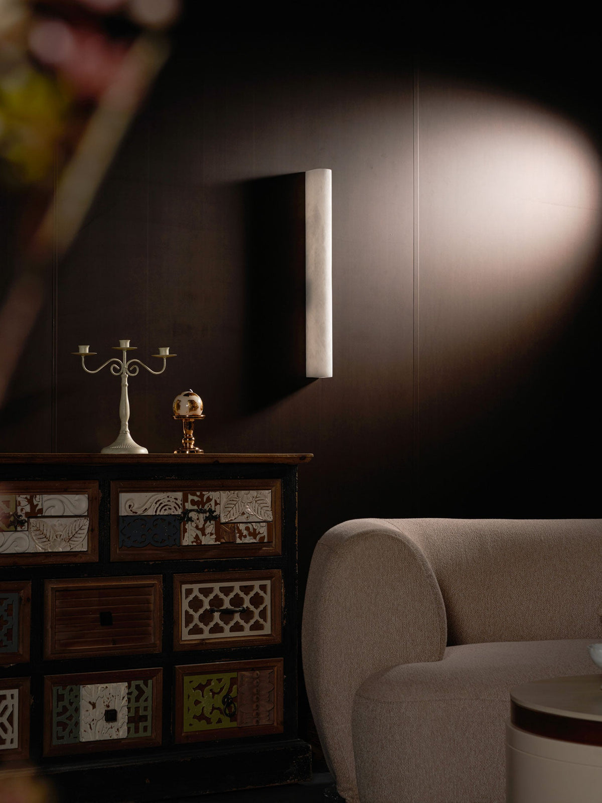 Unlit alabaster sconce on a dark wall lit by a soft spotlight, above a cabinet and a rounded cream armchair; from Clérachic.