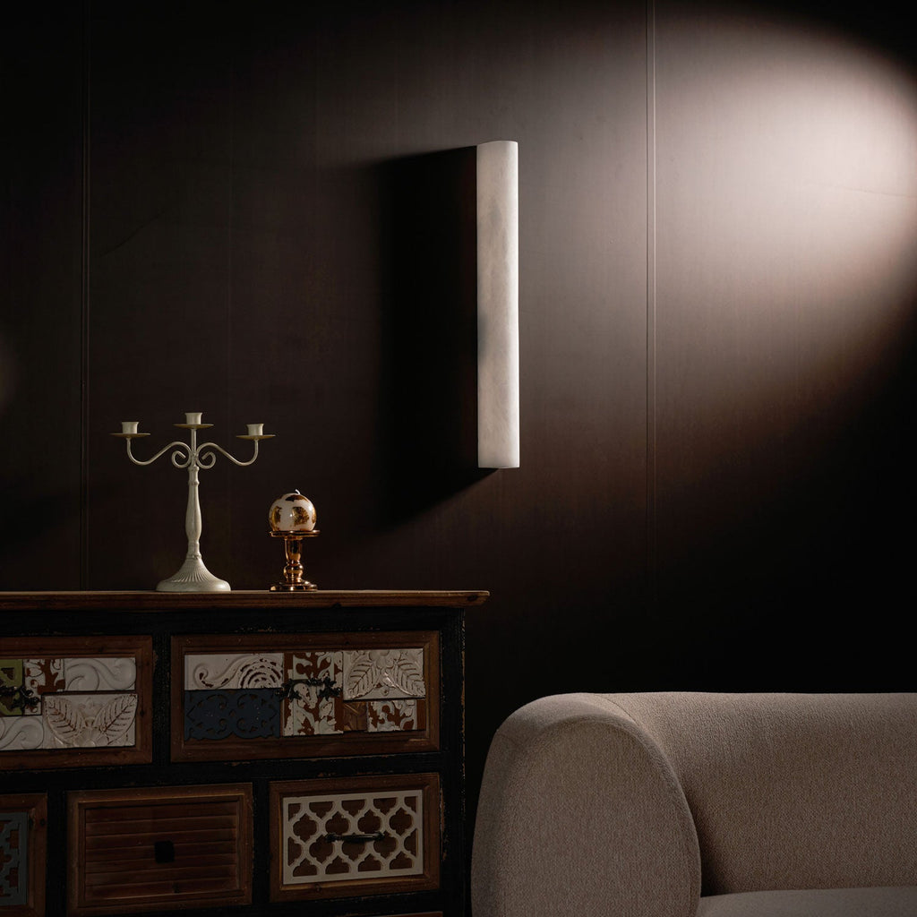 A pale Clérachic alabaster sconce, unlit, on a dark brown wall with a candelabra, a small globe and a cabinet below.