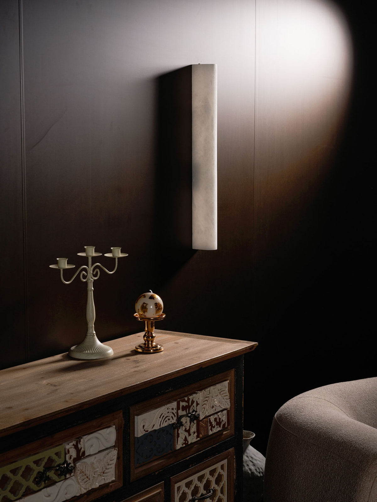 Angled view of the unlit Clérachic sconce on a dark wall, a candelabra and a copper-footed globe on the cabinet below.