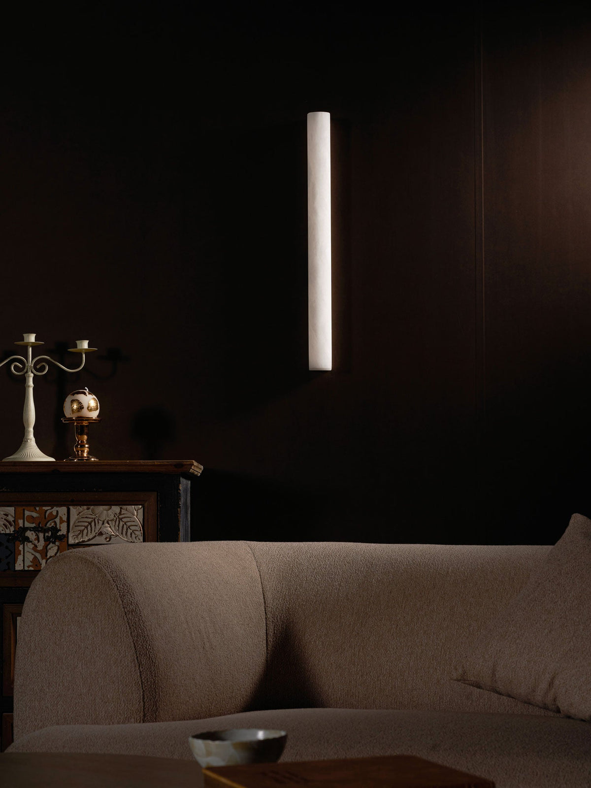 Plain white alabaster sconce, unlit, on a dark wall above a rounded sofa and a coffee table edge; this Clérachic wall light.