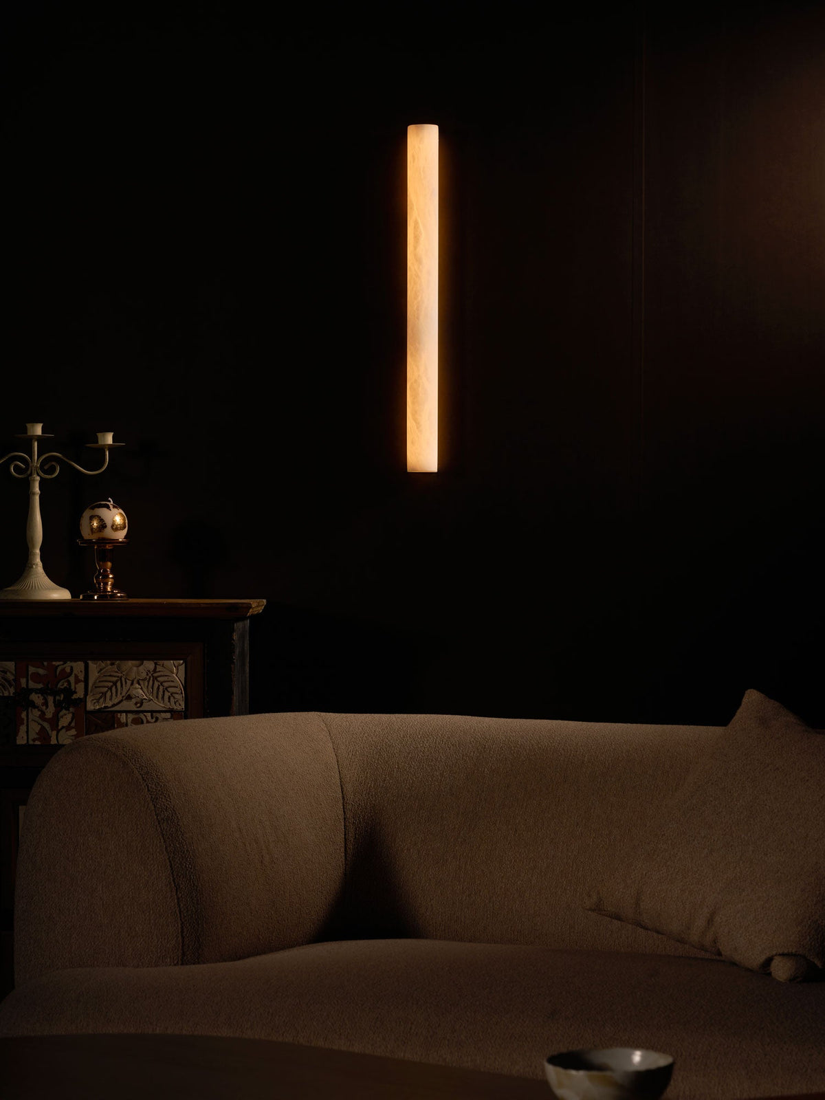 Tall amber glow from a Clérachic sconce on a dark wall, above a sofa and a cabinet with a candelabra at the left edge.