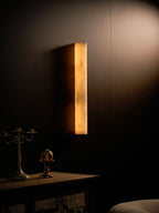 Close view of the lit Clérachic sconce, honey-amber alabaster glowing on a dark wall above a candelabra and a patterned cabinet.