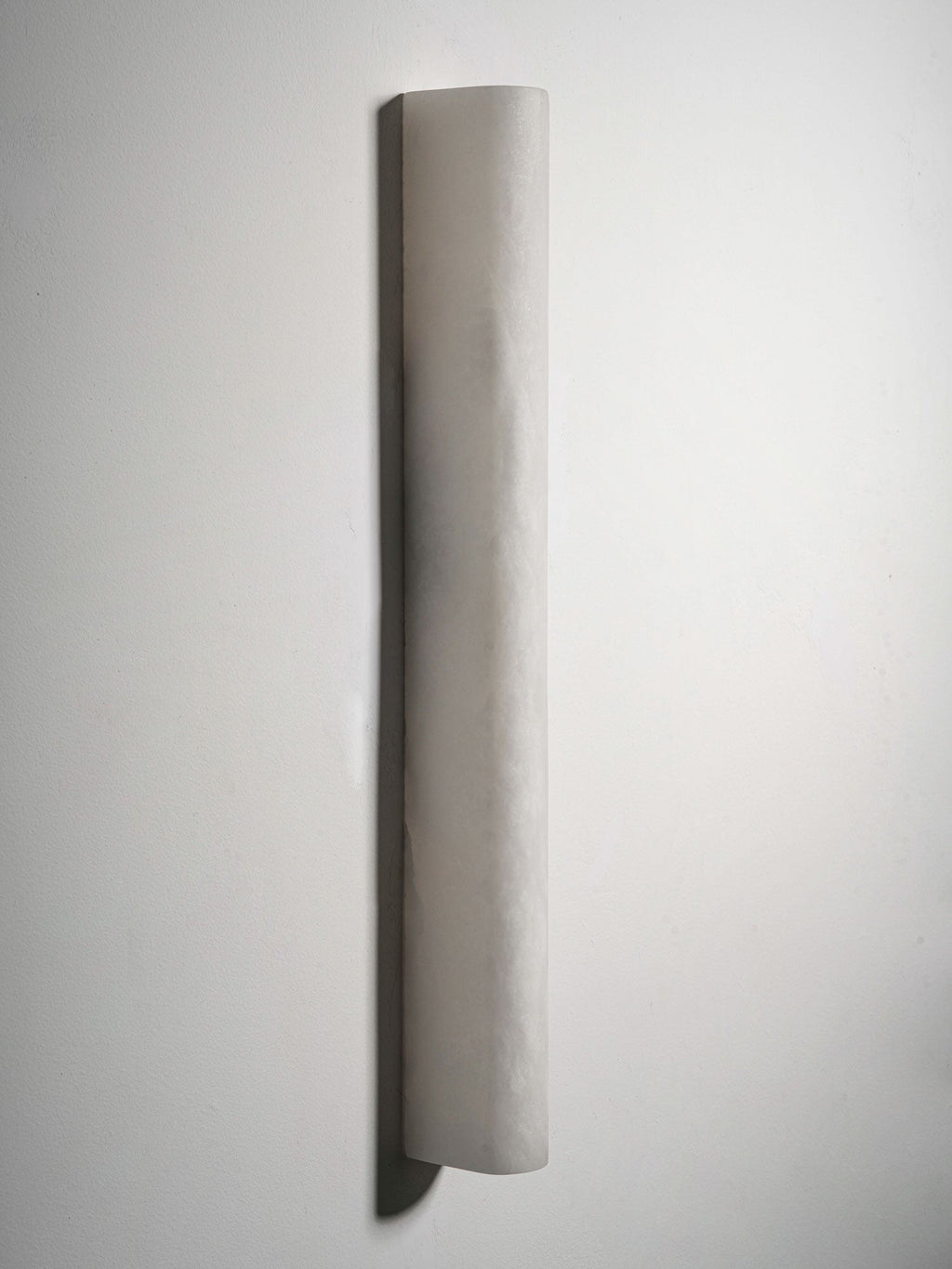 Wide Clérachic sconce shown unlit at a slight angle on a white wall, with a deep shadow along the left curve and a crinkled stone texture.