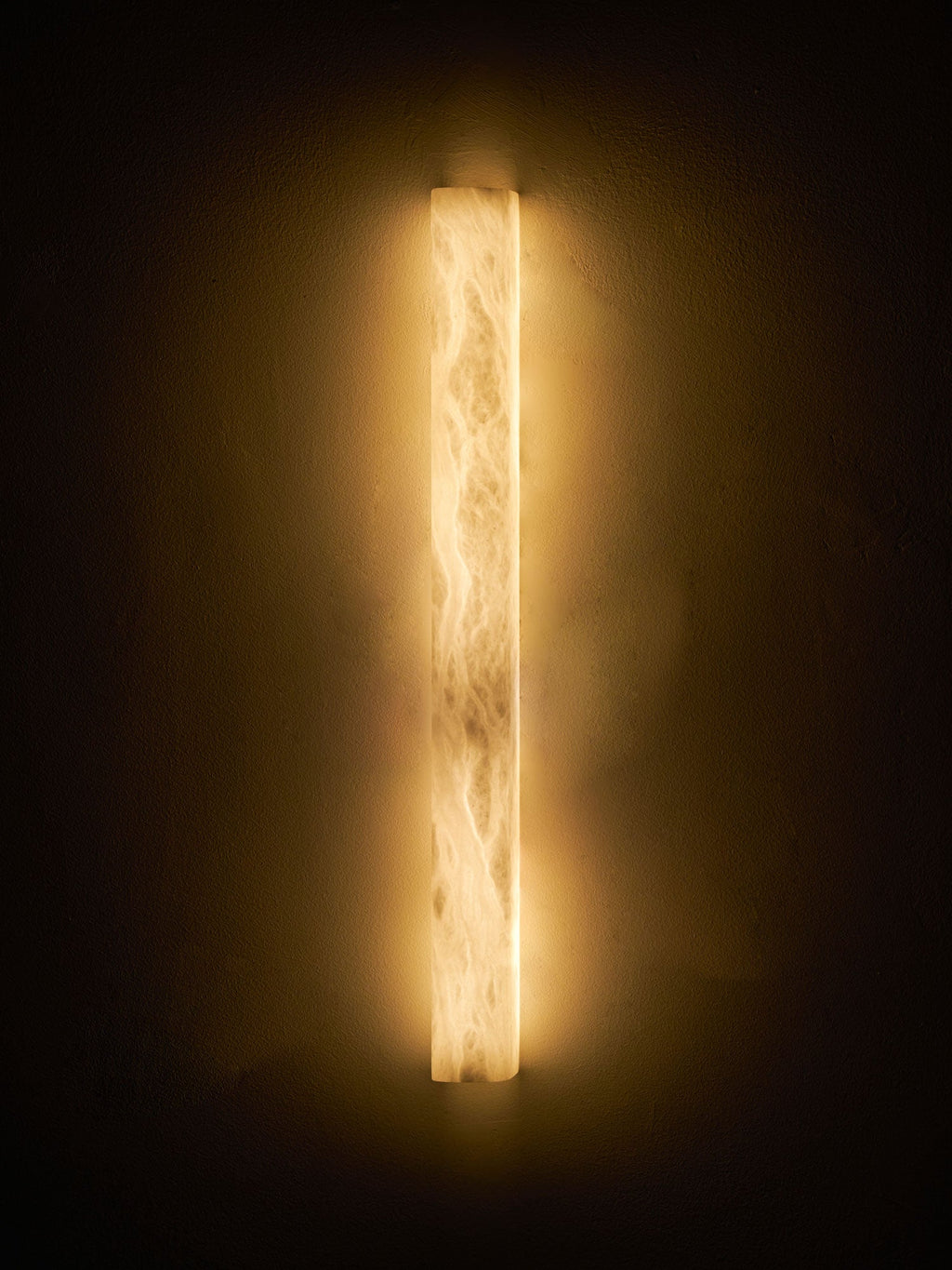 Slim alabaster sconce glowing warm on a dark wall, brightest at the top and bottom ends with cloudy veins between; from Clérachic.