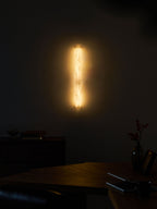 Distant view of the glowing sconce on a dark wall above a wooden table, a vase with berries and a notebook; a Clérachic wall light.