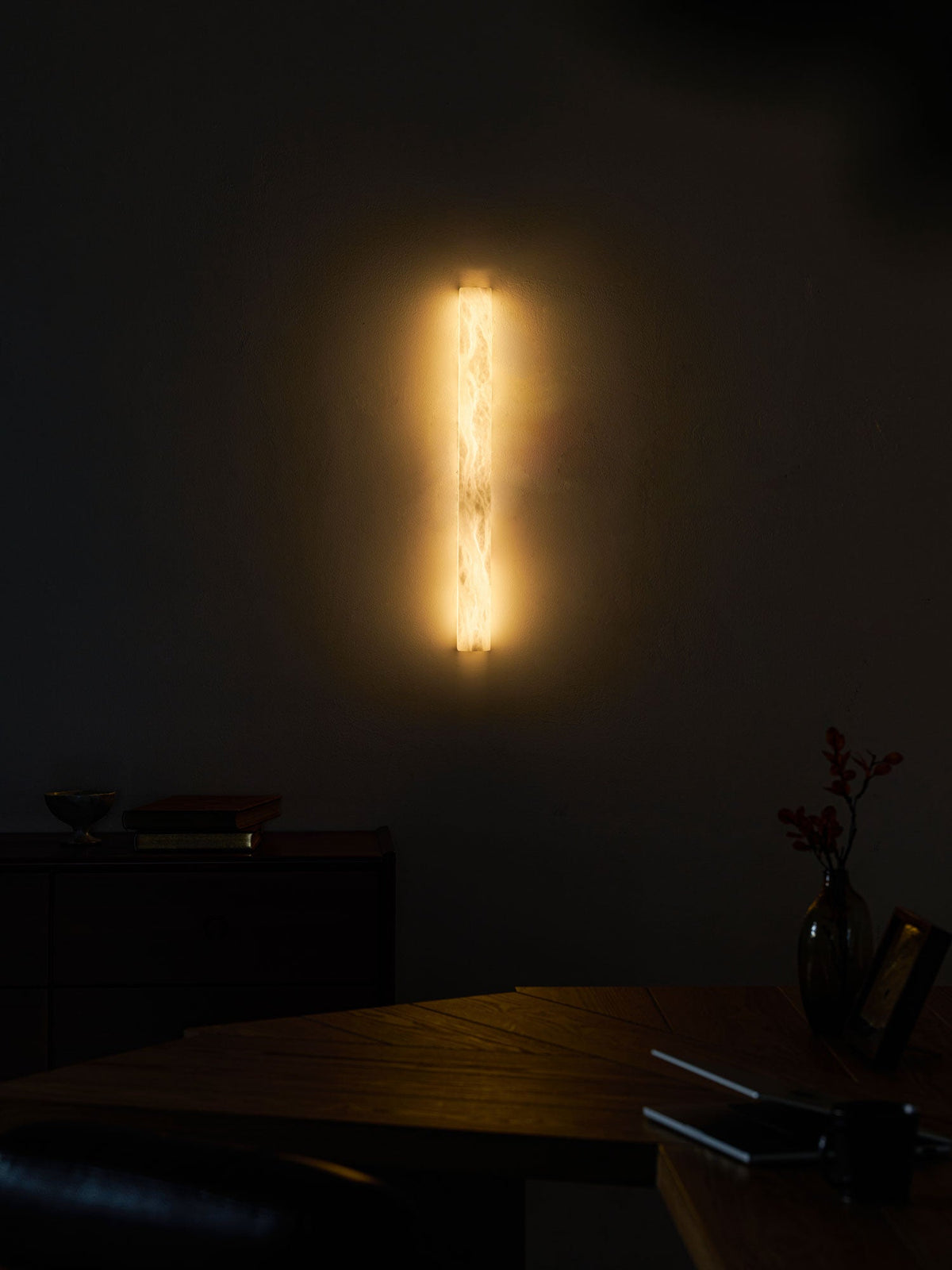 Distant view of the glowing sconce on a dark wall above a wooden table, a vase with berries and a notebook; a Clérachic wall light.