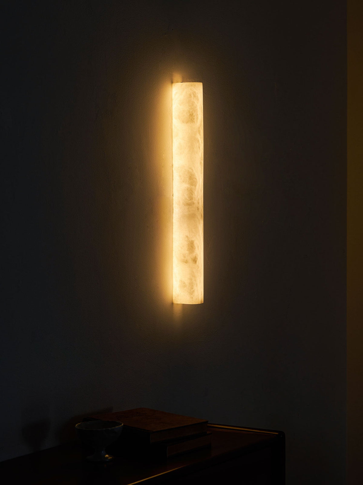 Lit Clérachic alabaster sconce on a dark wall above a low cabinet, with books and a glass bowl below in the dim light.