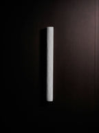 Unlit slim Clérachic sconce in pale stone on a dark brown wall, a narrow vertical bar with a soft, rough surface.